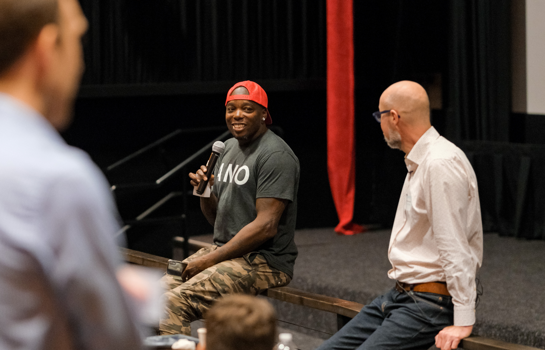 Man looks on as other man speaks into a microphone